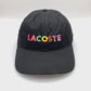 Spinning Black Lacoste 6 Panel Hat with multi colored Lacoste letters on the front panel, and white background.