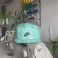 The making of a green NIKE Swim Shorts upcycled into a 5 Panel Hat, black iconic NIKE logo in front, with a white background.
